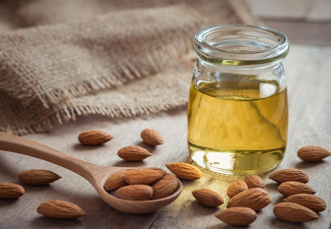 Almond oil in glass jar surrounded by whole almonds on wooden spoon.