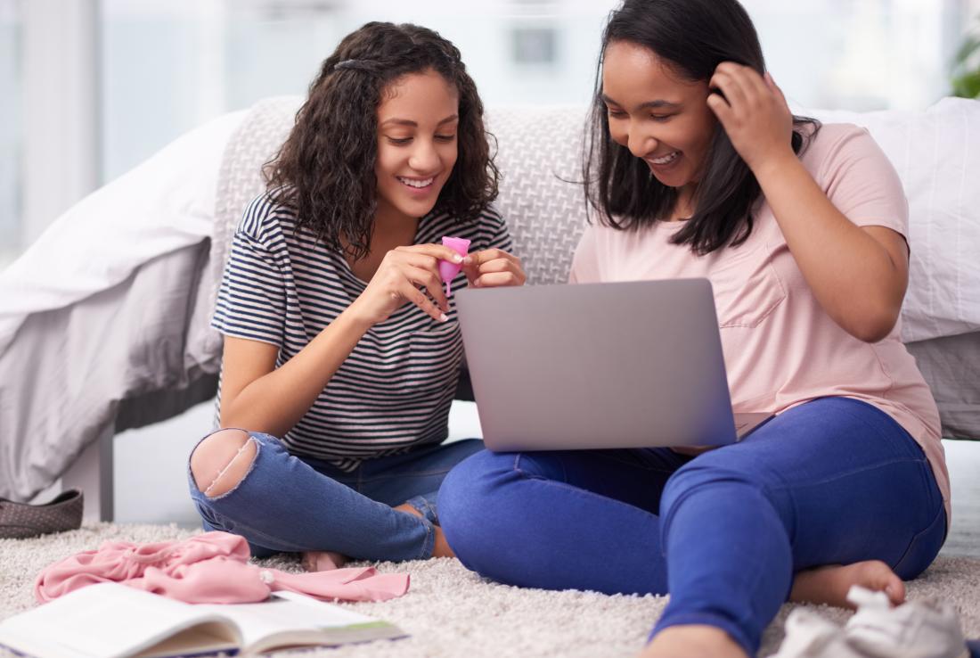 Two girls sitting on bed holding menstrual cup looking at laptop
