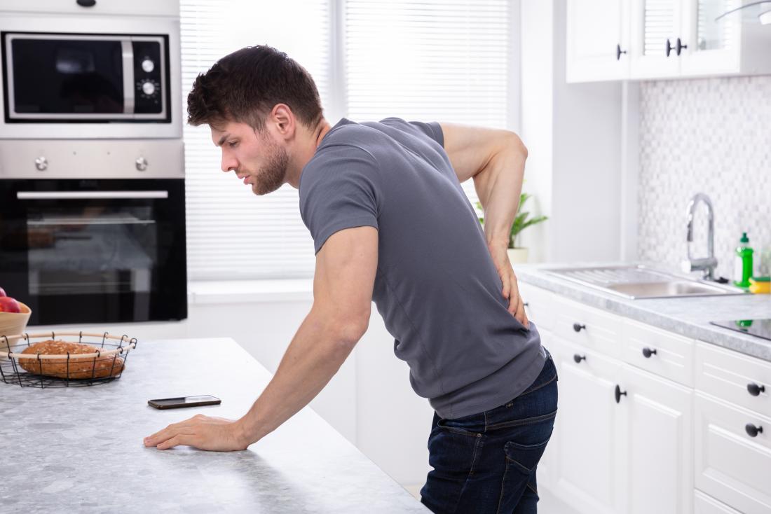 A man with back and side pain due to a UTI that is causing blood in the urine leans on the kitchen counter.