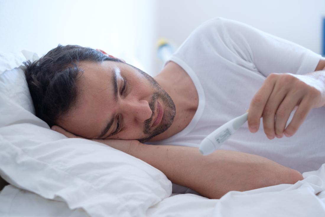 man with fever temperature lying in bed looking at thermometer