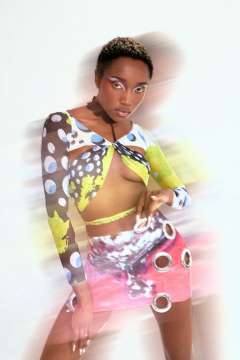 a black woman with short hair wears a chartreuse, white, blue, and black cropped top and pink patterned skirt with grommets