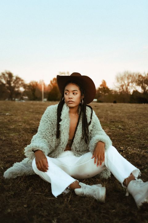 megan wilson barrel racer in a brown cowboy hat and fuzzy jacket with white jeans sitting on grass