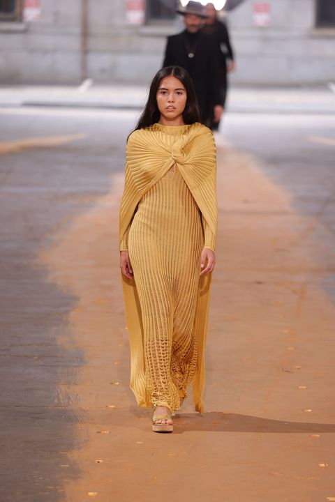 a brunette woman wearing a gold column gown walks down a runway