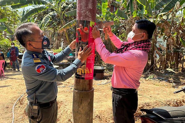 A man in a mask, wearing a uniform with patches on the shoulders and carrying a walkie-talkie, is holding up a poster around a telephone pole. Another man, wearing a pink long-sleeve shirt and a pink checkered scarf, is hammering the poster into the pole. A villager is standing behind them next to a wall of tropical vegetation.