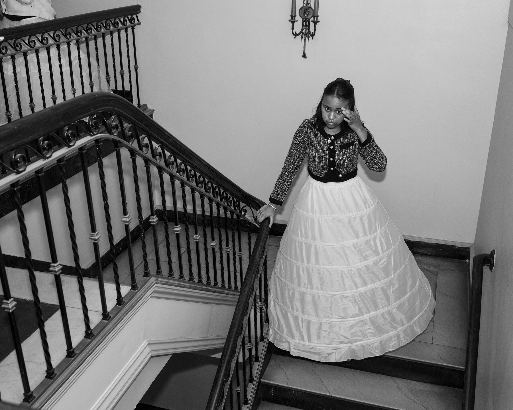 A young woman wearing a white hoop skirt and a plaid cardigan sweater with black trim standing halfway down a staircase with an ornate, black metal rail. She holds the bannister with her right hand and touches her eyebrow with her left.