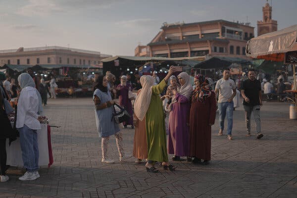 A group of women in long, colorful, traditional Moroccan robes and head coverings, congregate in a plaza with a surface of geometrically arranged pale brown tiles. There are covered market stalls in the background, more people and various buildings.