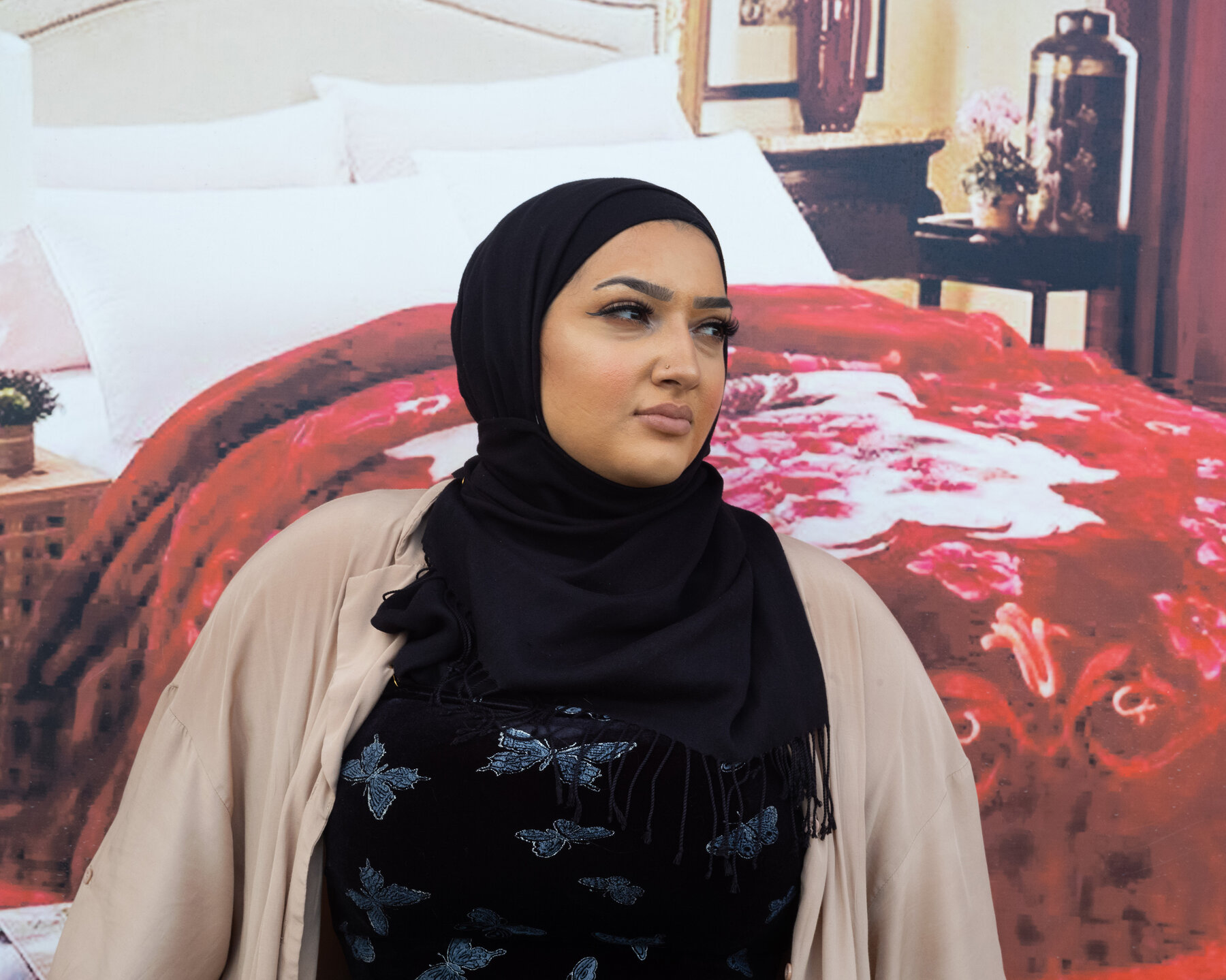 A young woman standing in front of an advertisement depicting a bedroom with a bed covered with a red floral blanket. She wears a black shirt covered in blue butterflies, a tan cardigan sweater and a black headscarf.