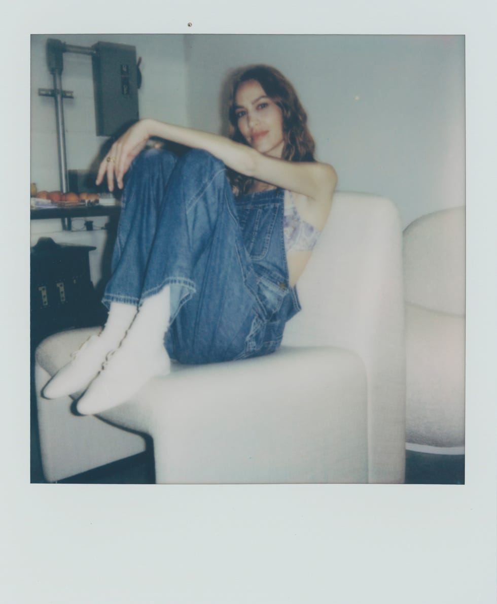 a woman in denim overalls and white sneakers sitting on a white chair