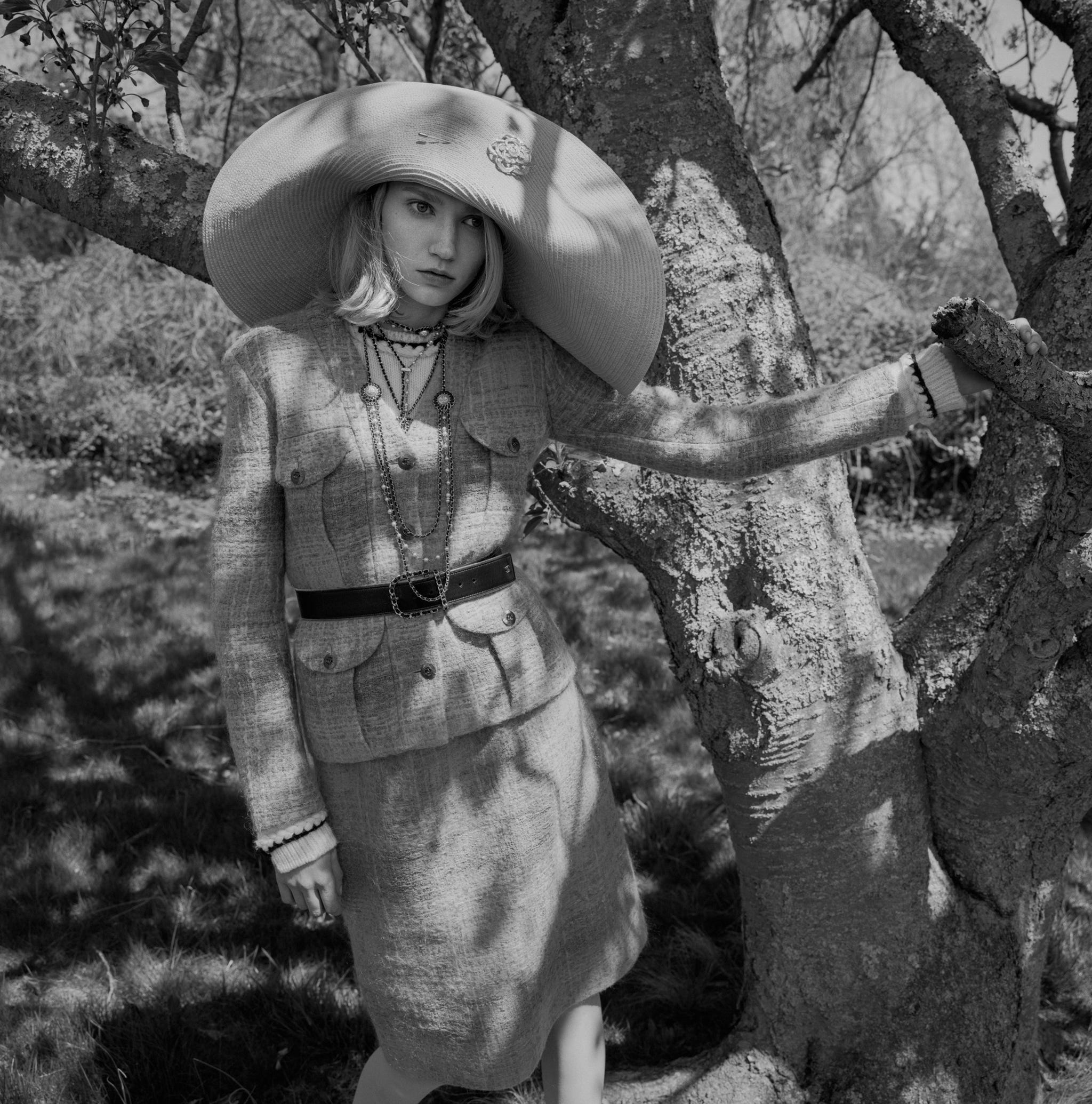 a black and white photo of a woman in a tweed suit and an oversized hat standing next to trees