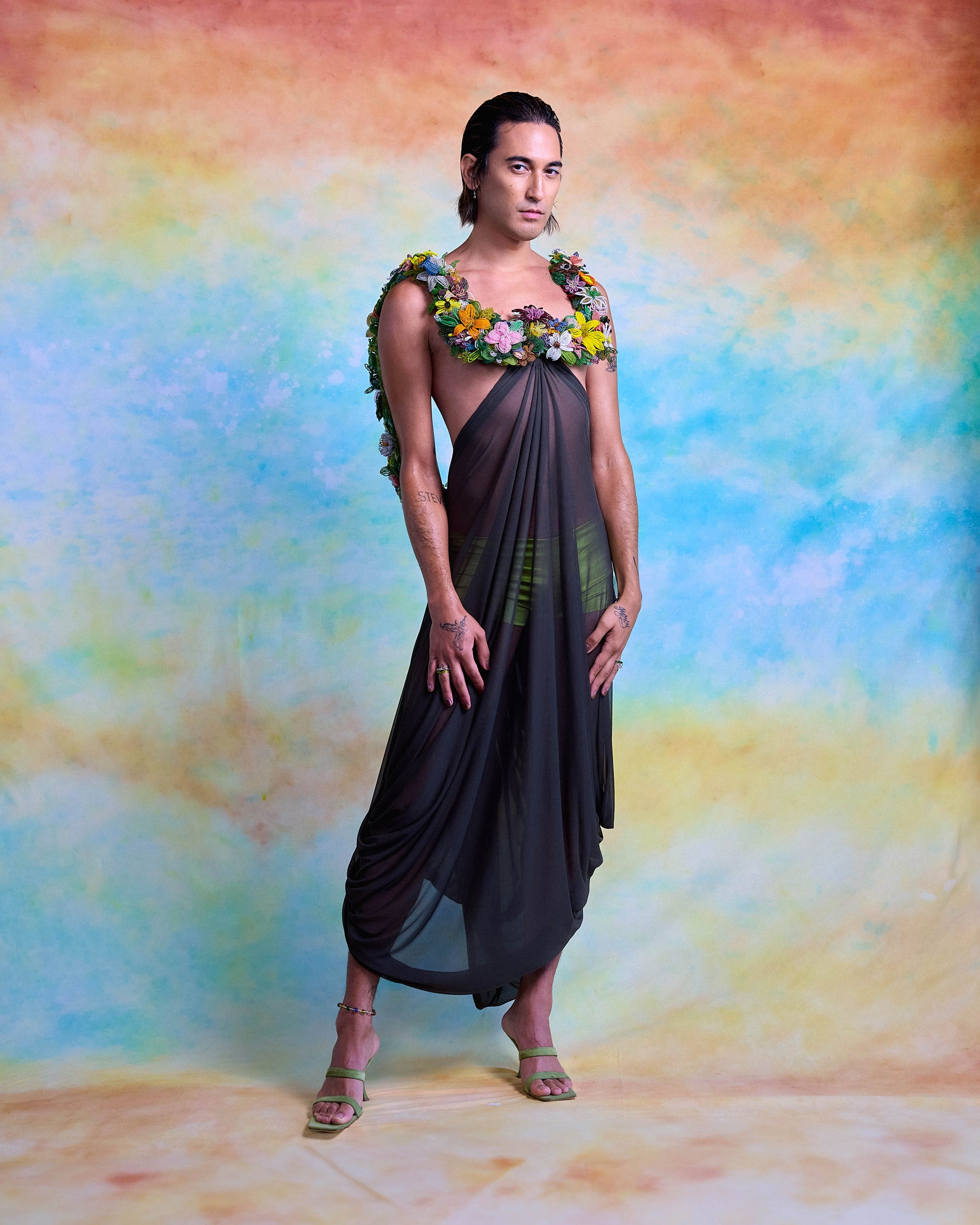 a model stands in a striking pose, wearing a dark, draped garment adorned with colorful flowers around the neckline