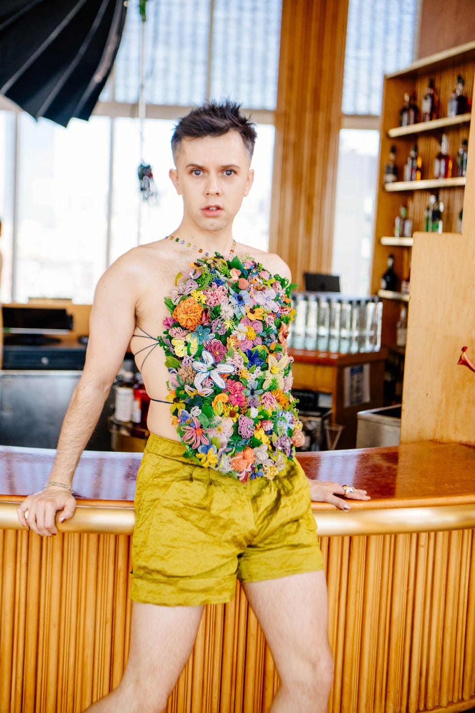 a figure stands confidently at a bar, wearing a vibrant chest piece made of numerous colorful flowers