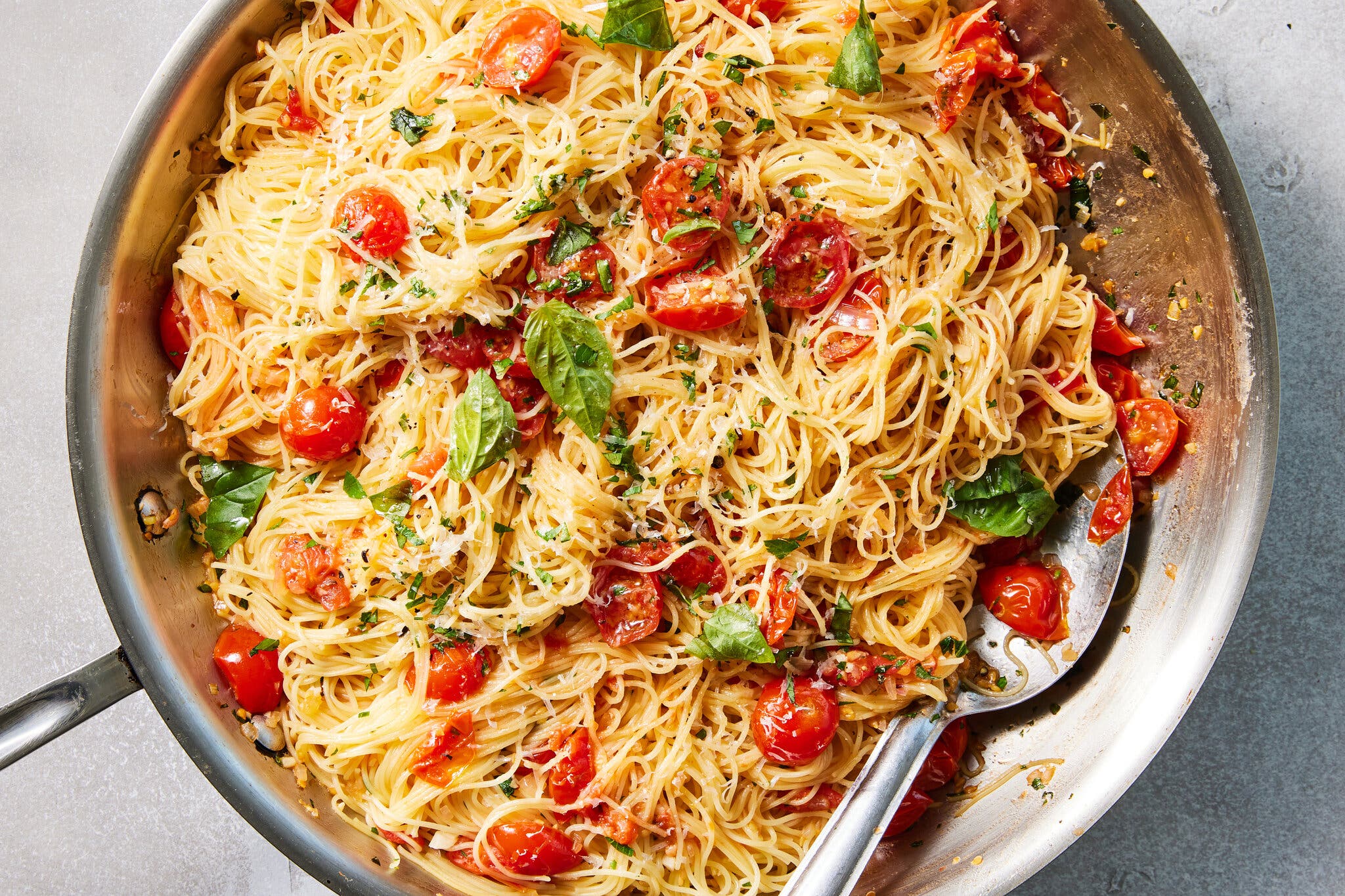 A stainless steel skillet on a gray background is full of thin noodles tossed with burst cherry tomatoes and basil leaves.