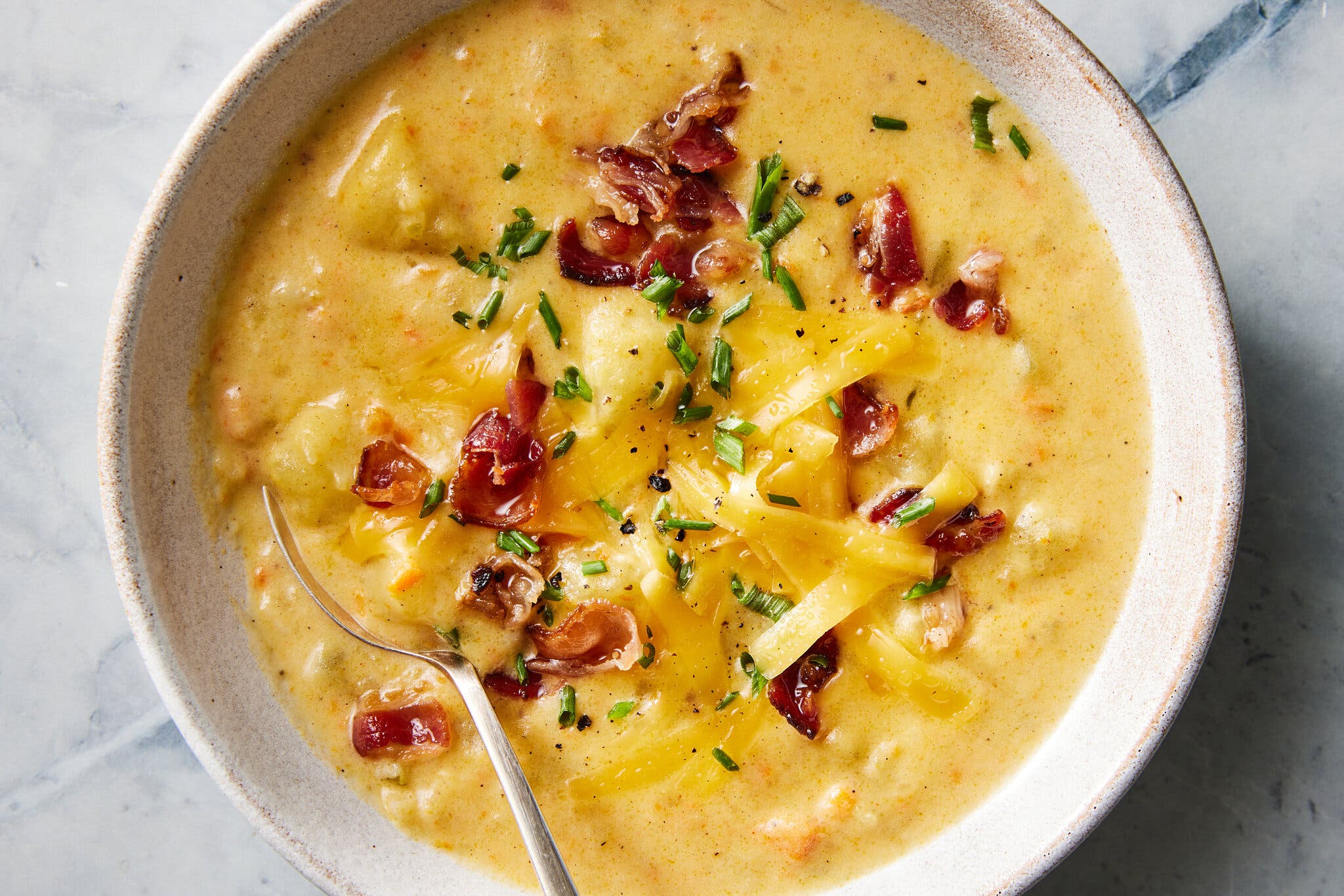 A white ceramic bowl holds potato soup topped with shredded cheese, crumbled bacon and snipped chives.