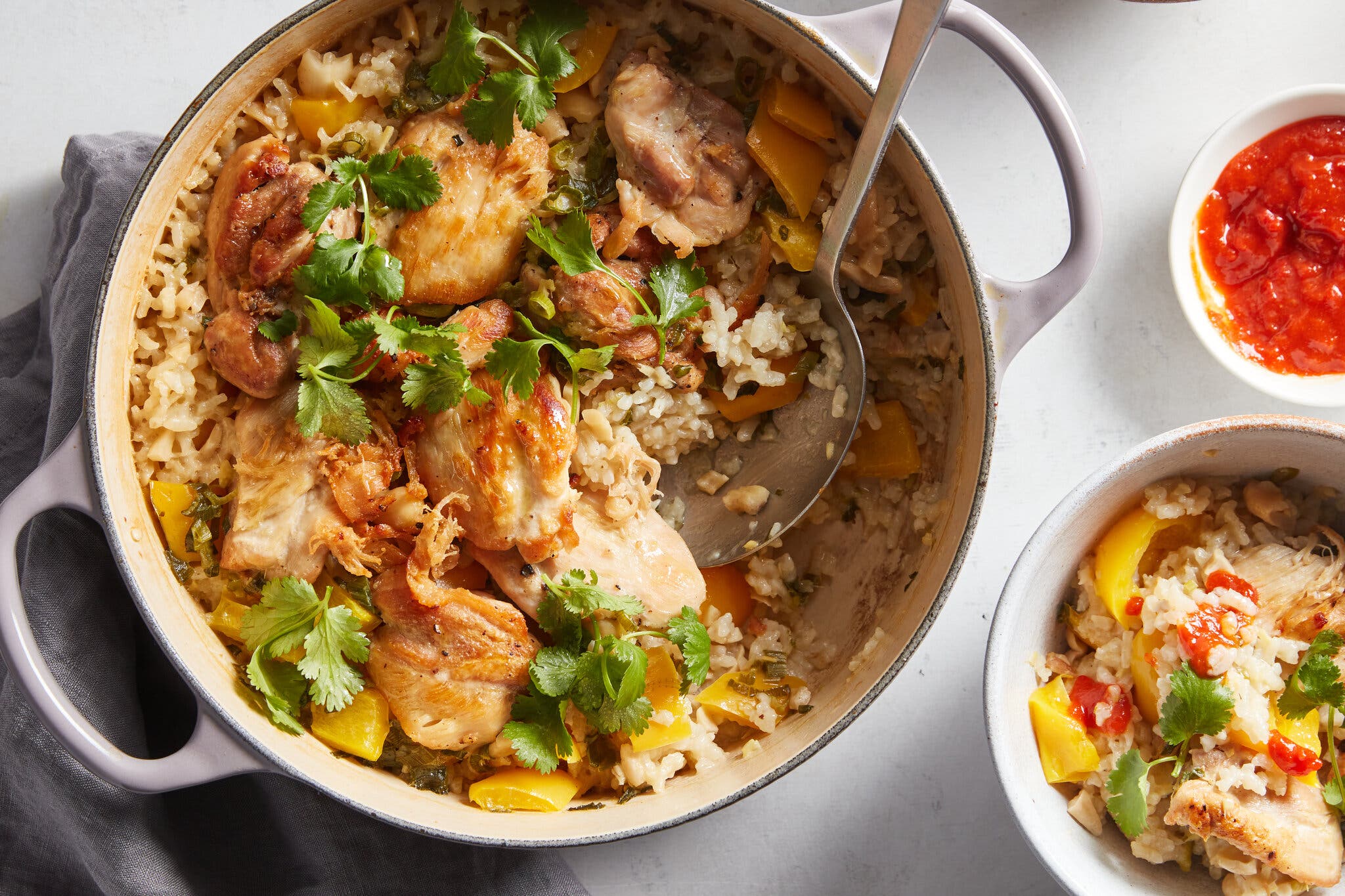 A Dutch oven holds sticky coconut chicken and rice with a serving scooped out. A small bowl of hot sauce is nearby.