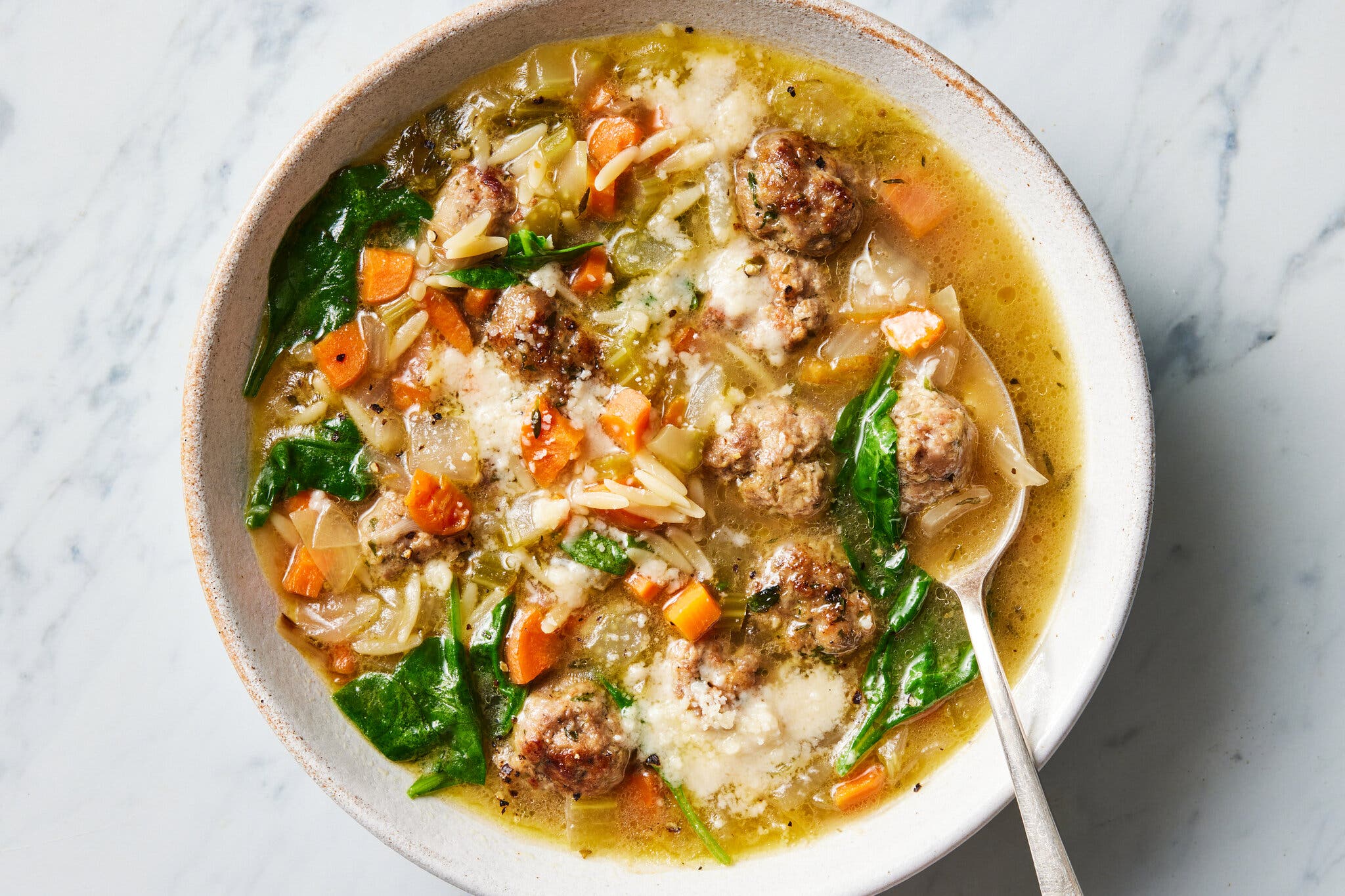 An overhead image of a white bowl filled with light brown meatballs, orzo pasta, carrot cubes, wilted spinach in a yellow broth. Everything is dusted with a melty layer of grated Parmesan. 