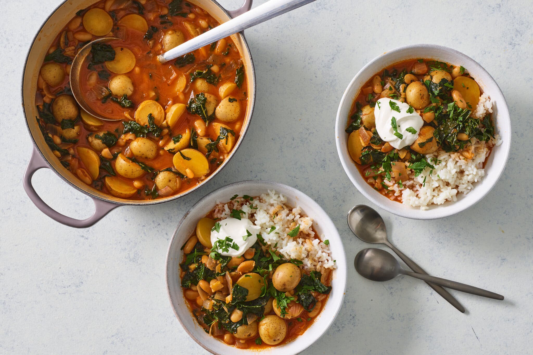 A Dutch oven holds gochujang potato stew with two servings in bowls; the servings also have white rice and sour cream.