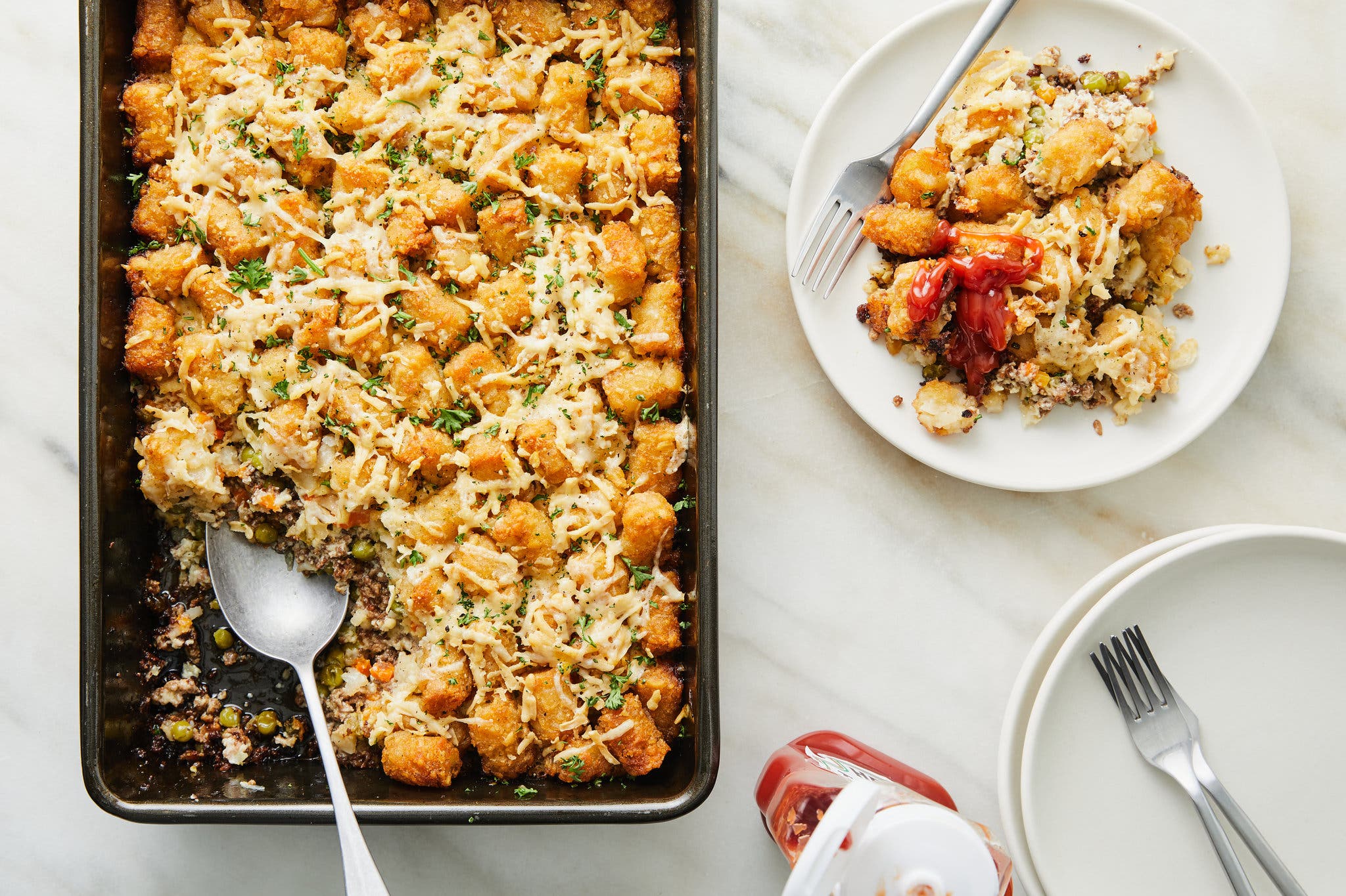 An overhead image of a black baking dish filled with tater tots atop a brown ground meat mixture with a spoonful removed. The entire casserole is sprinkled with melted shredded cheese. A single plate is off to the side with a serving on top. 