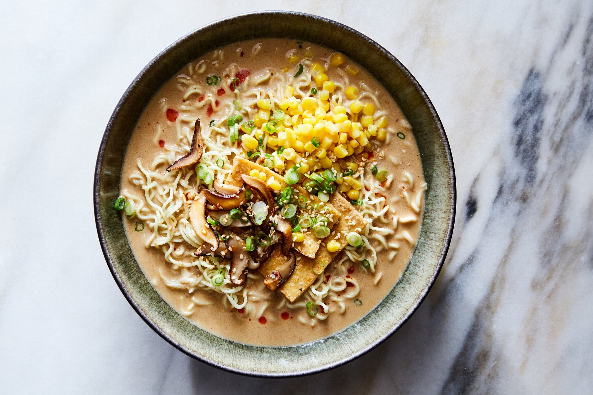 An overhead image of a bowl of tantanmen topped with tofu, mushrooms, corn and scallions.