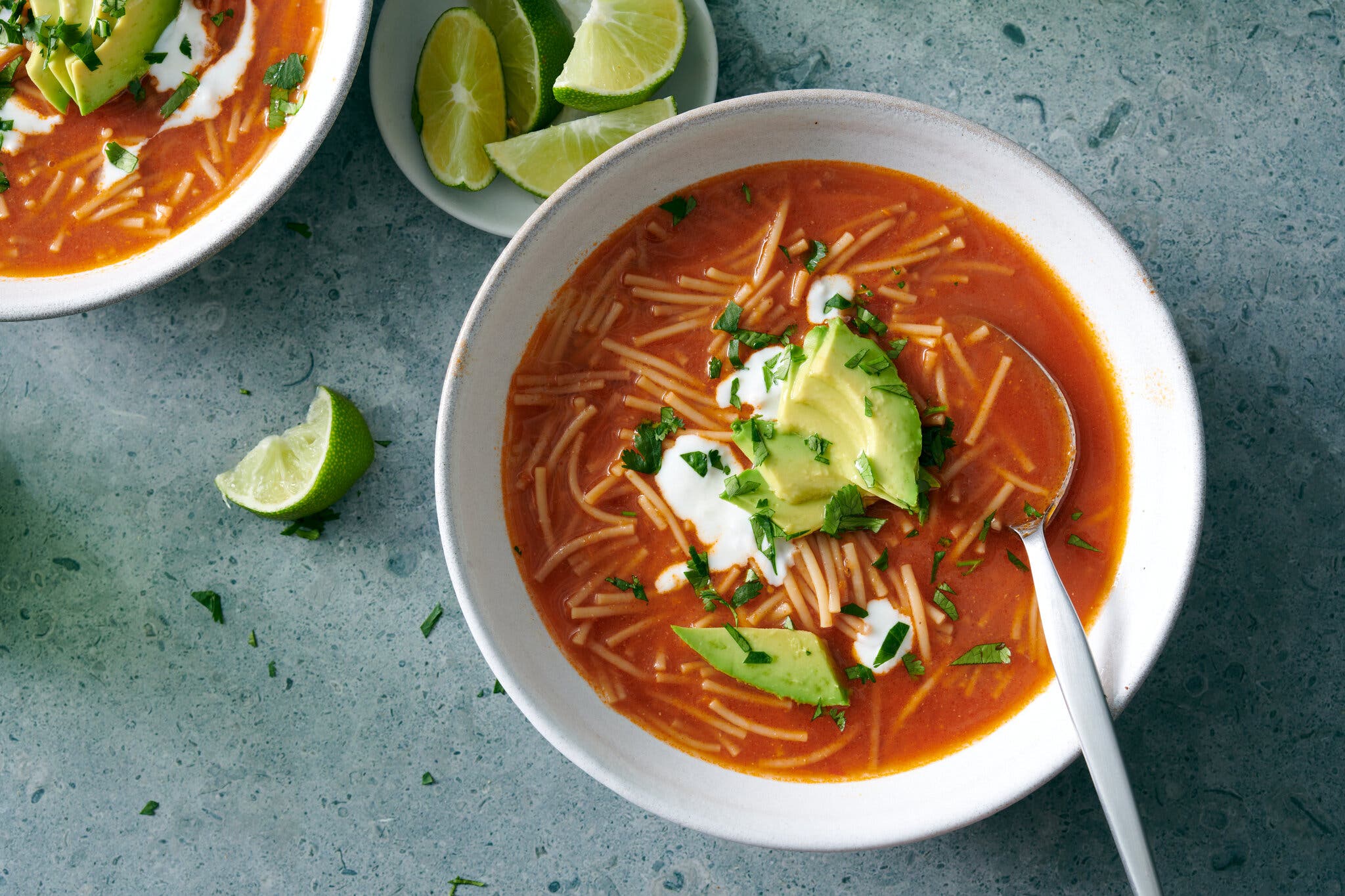 A bowl of noodle soup with a bright red broth is topped with cream, avocados and herbs. Lime wedges sit off to the side, as does another serving.