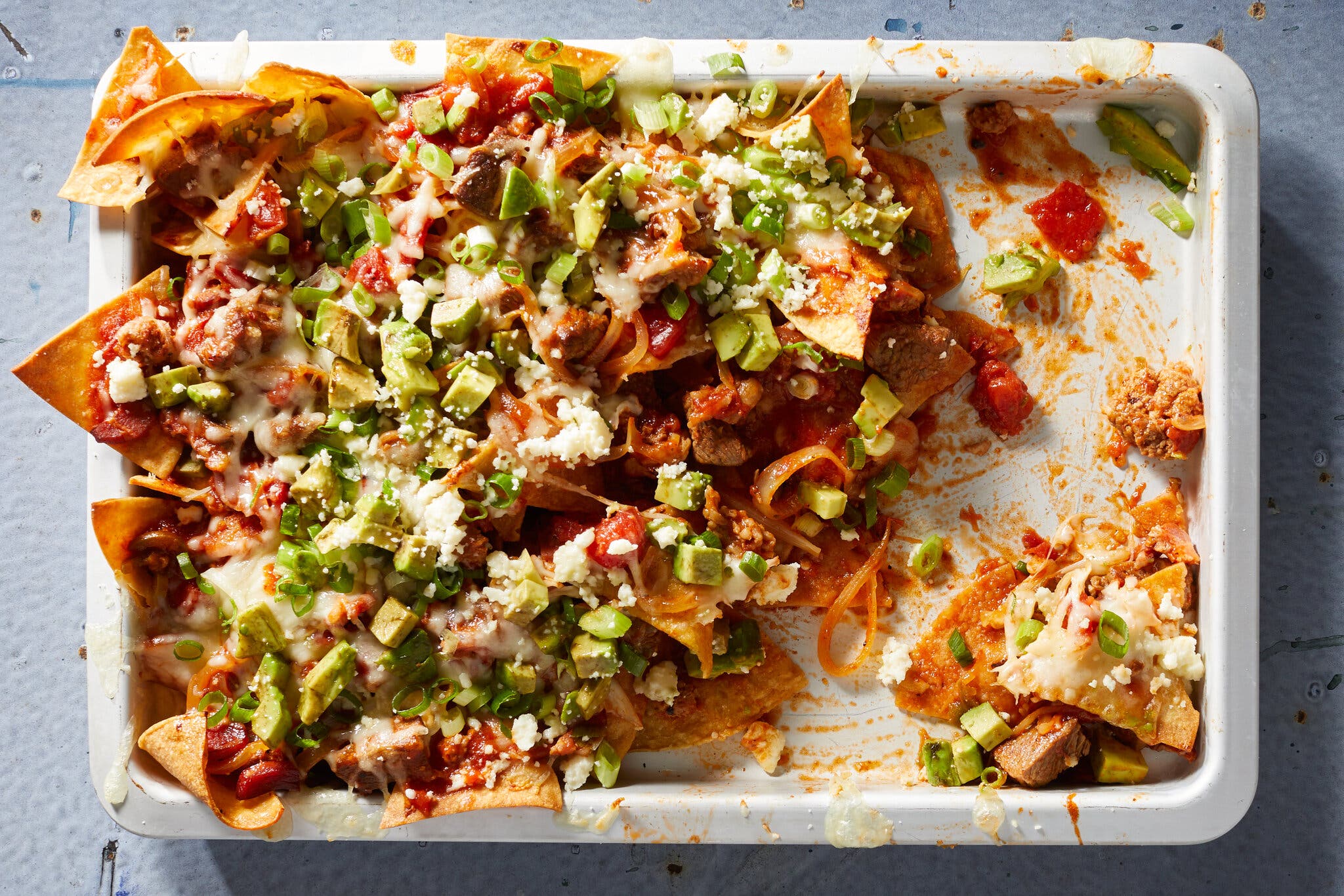 An overhead image of a big white pan partially filled with tortilla chips topped with melted cheese, chunks of beef, avocados, scallions and roasted tomato sauce. 