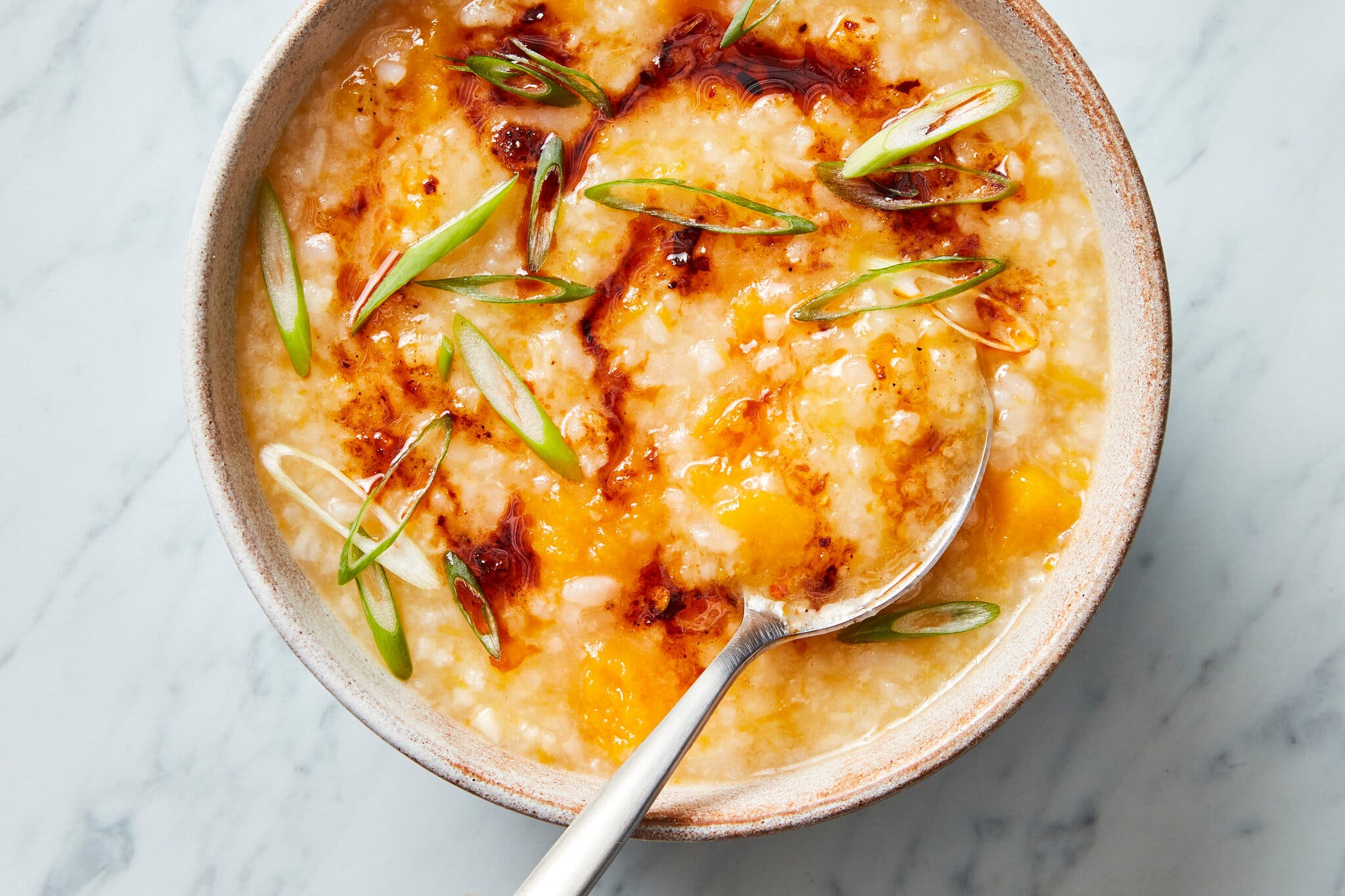 A tan bowl is filled with rice porridge studded with bits of butternut squash and topped with chile oil and scallions.