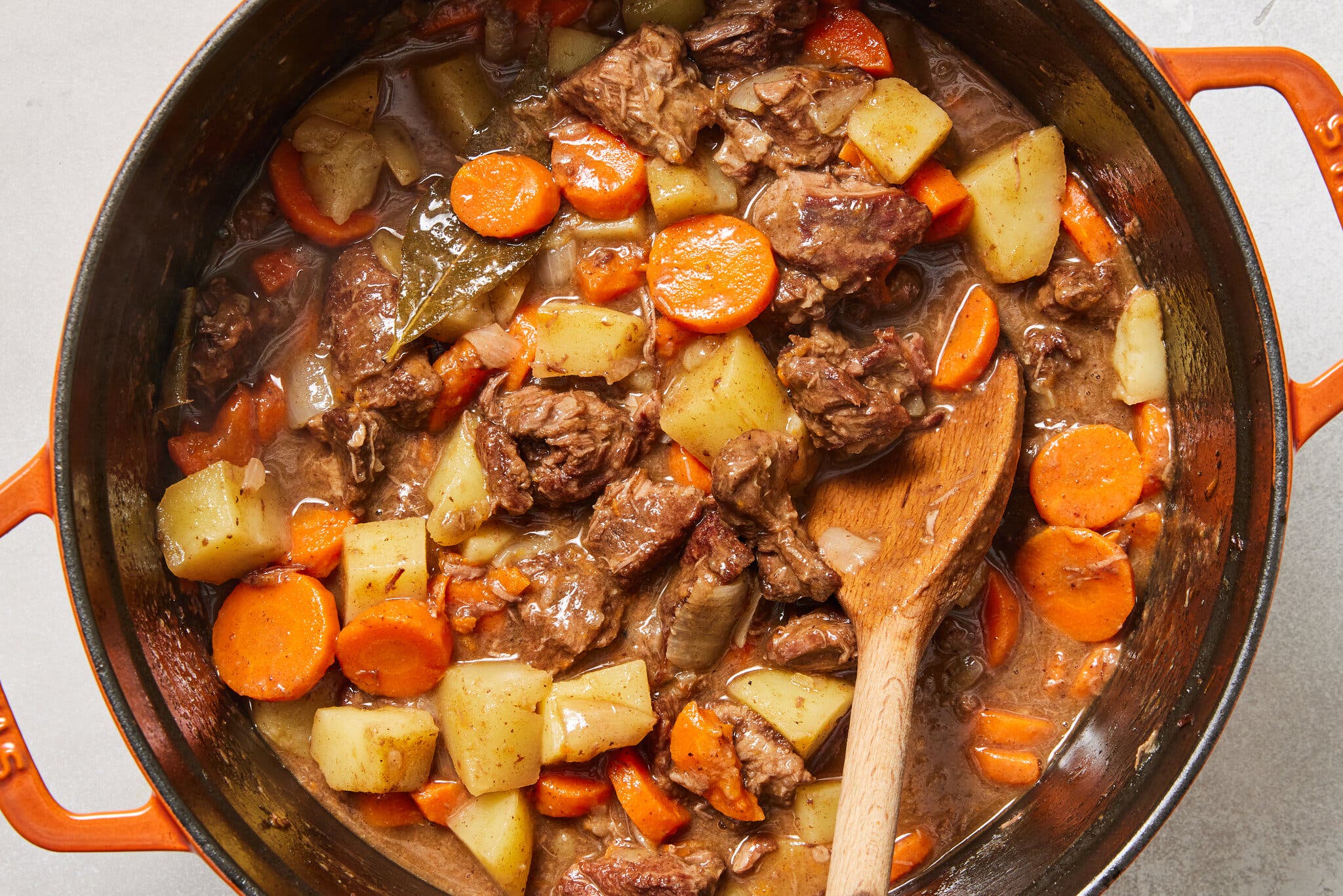 An overhead image of an orange cast-iron enamel two-handled pot filled with a thick and saucy mixture of beef chunks, potatoes cubes and carrot circles.