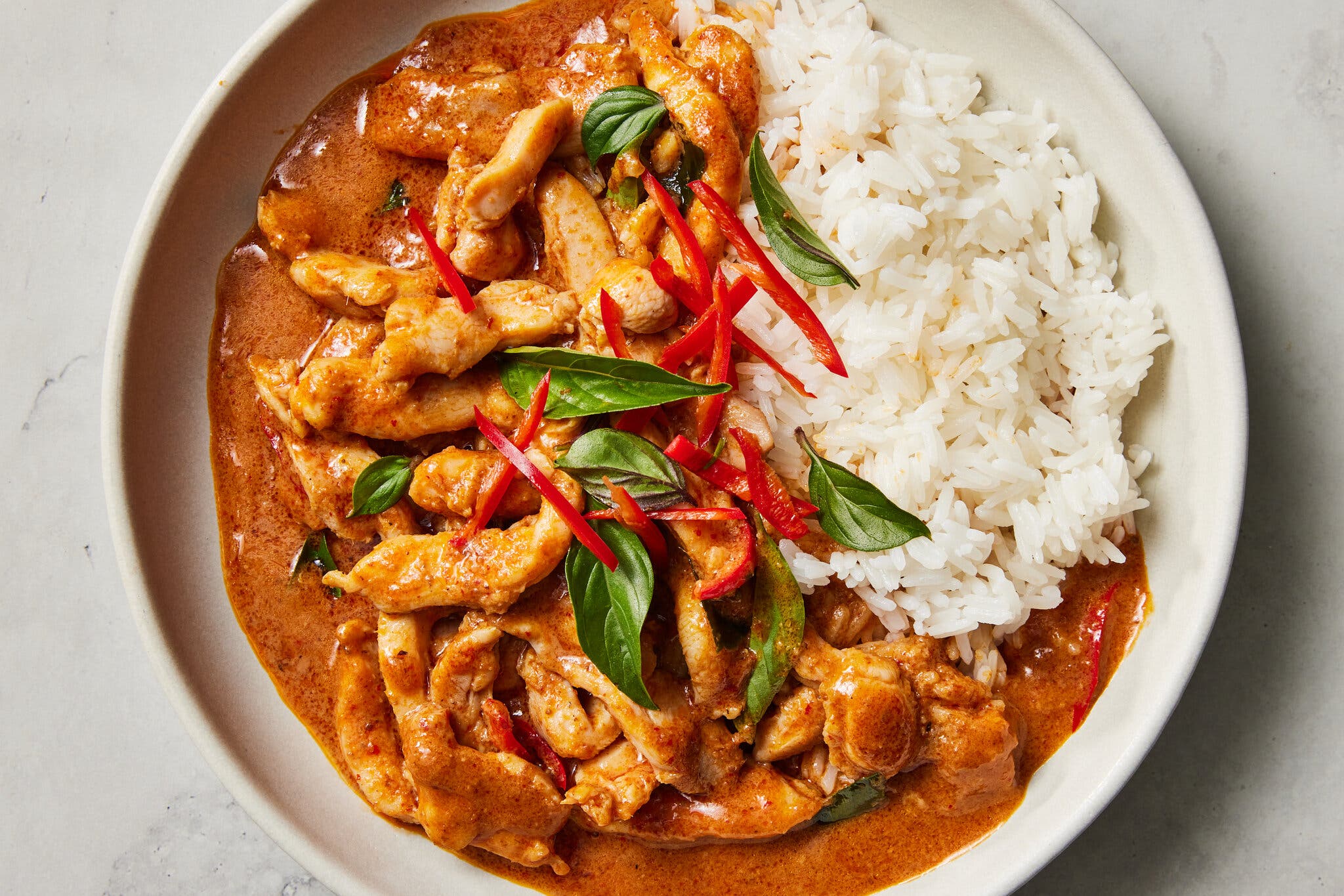 A shallow white bowl holds panang curry, topped with basil leaves and slivered red pepper, as well as steamed white rice.