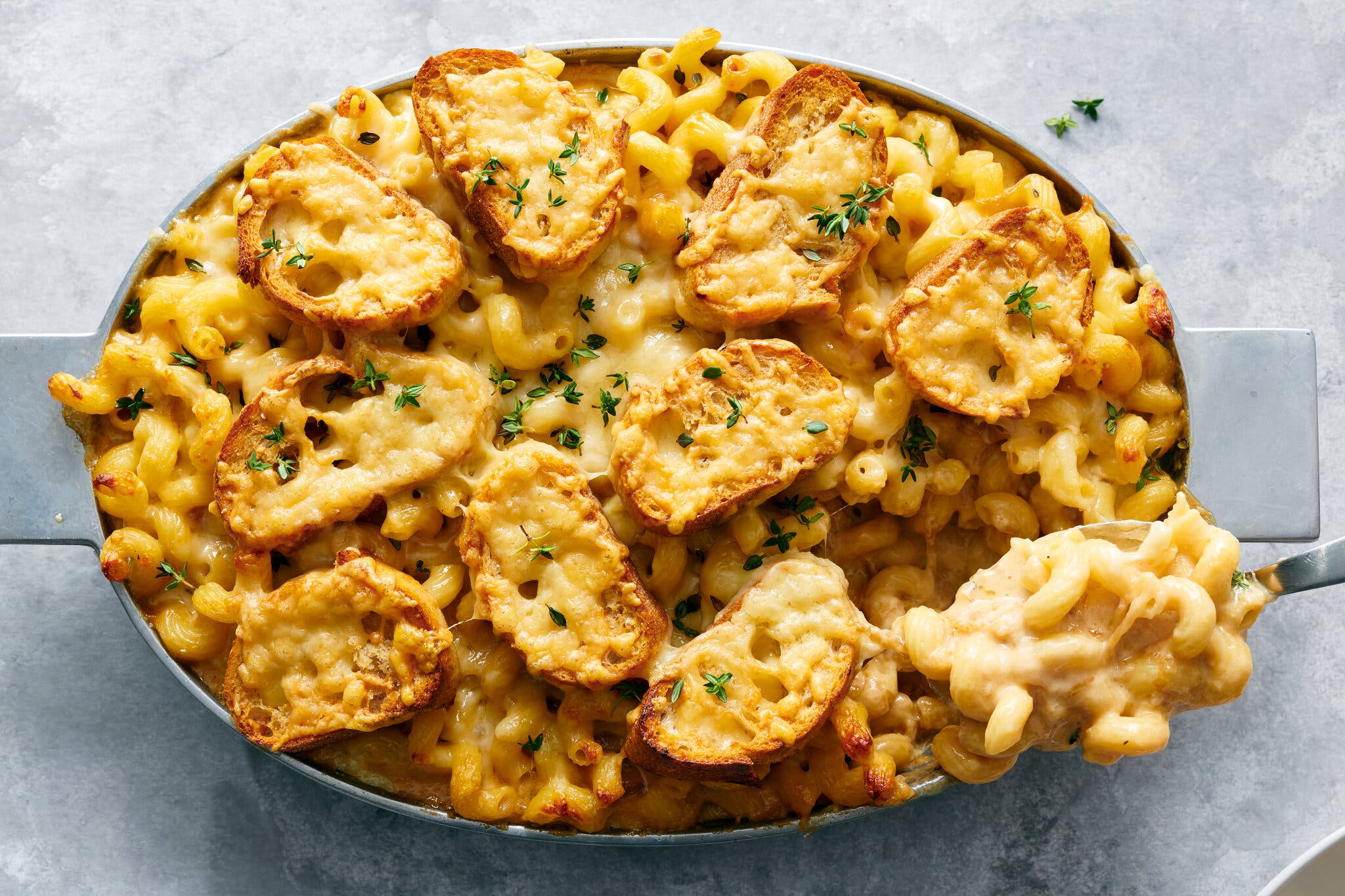 A silver dish is full of yellow curly pasta topped with slices of baguette covered in browned, melted cheese. A spoonful is being removed. Everything is sprinkled with thyme leaves. 