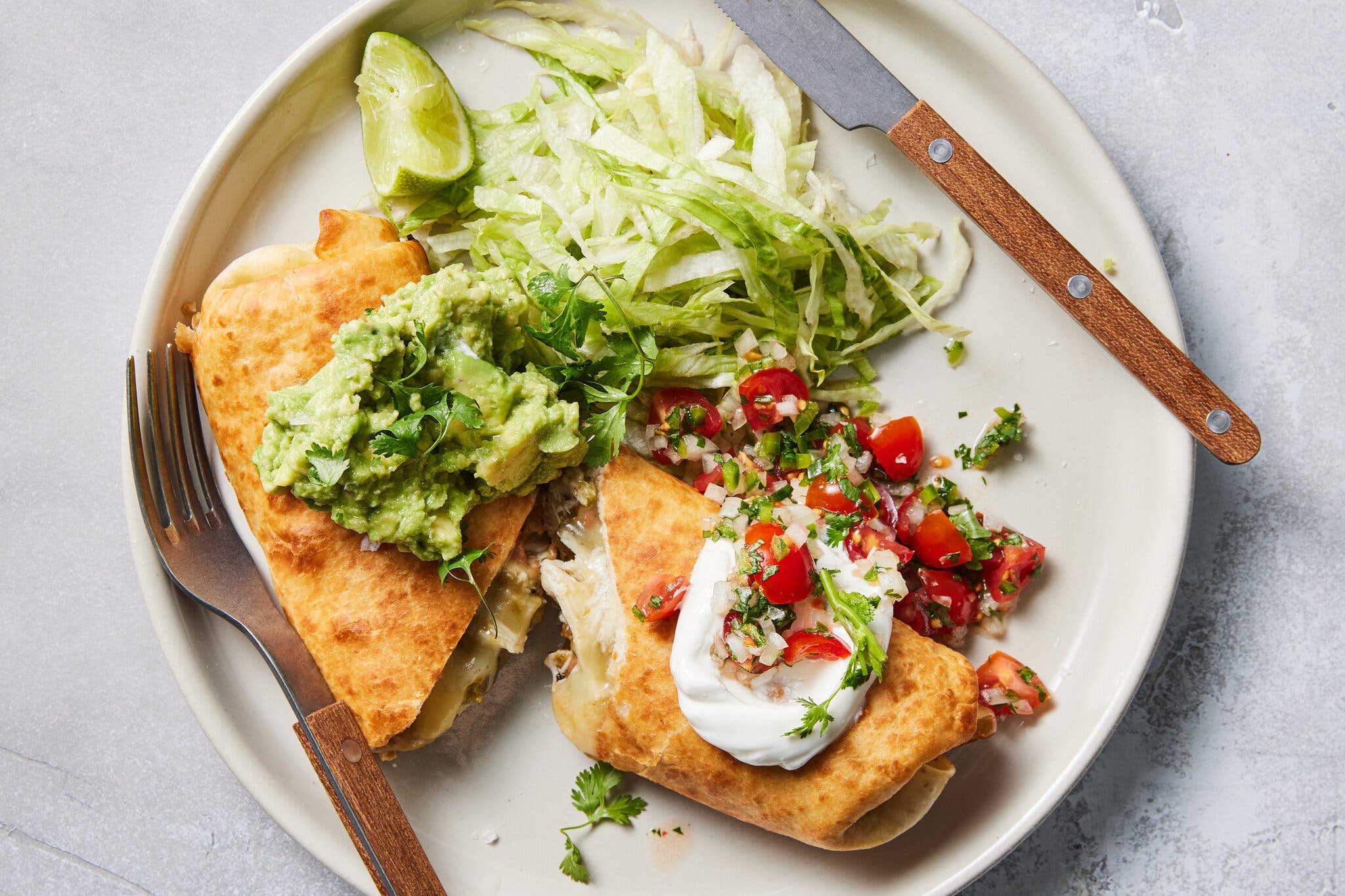 An overhead shot of a chimichanga cut open on a white plate topped with sour cream, guacamole, salsa and shredded lettuce.