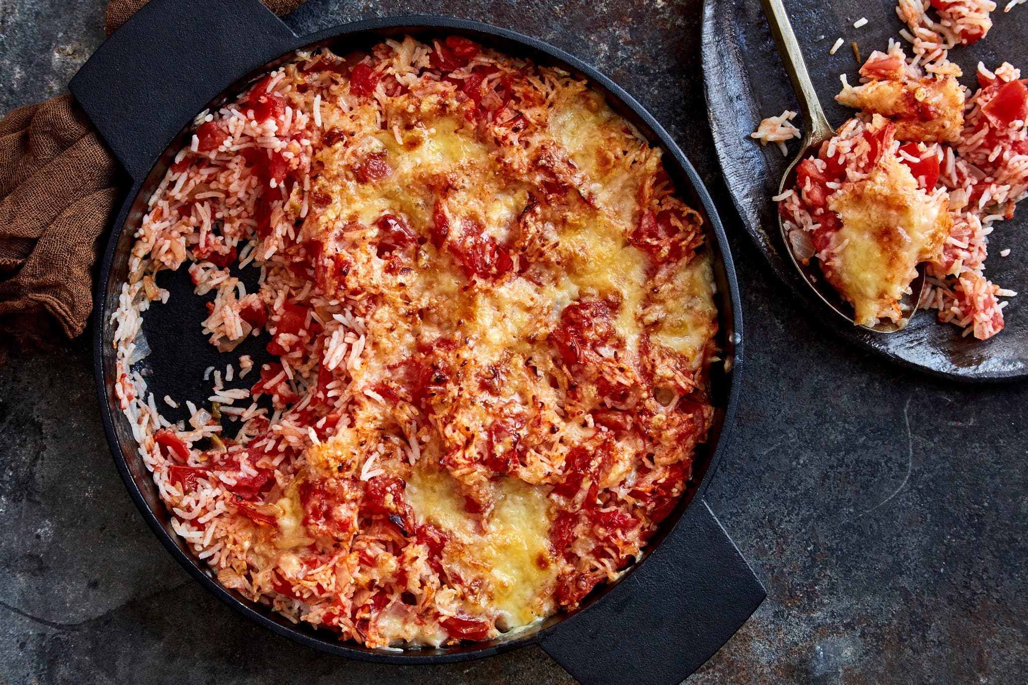 A cast-iron skillet is full of tomato rice with crispy Cheddar, a portion of which has been scooped out onto a dark plate.