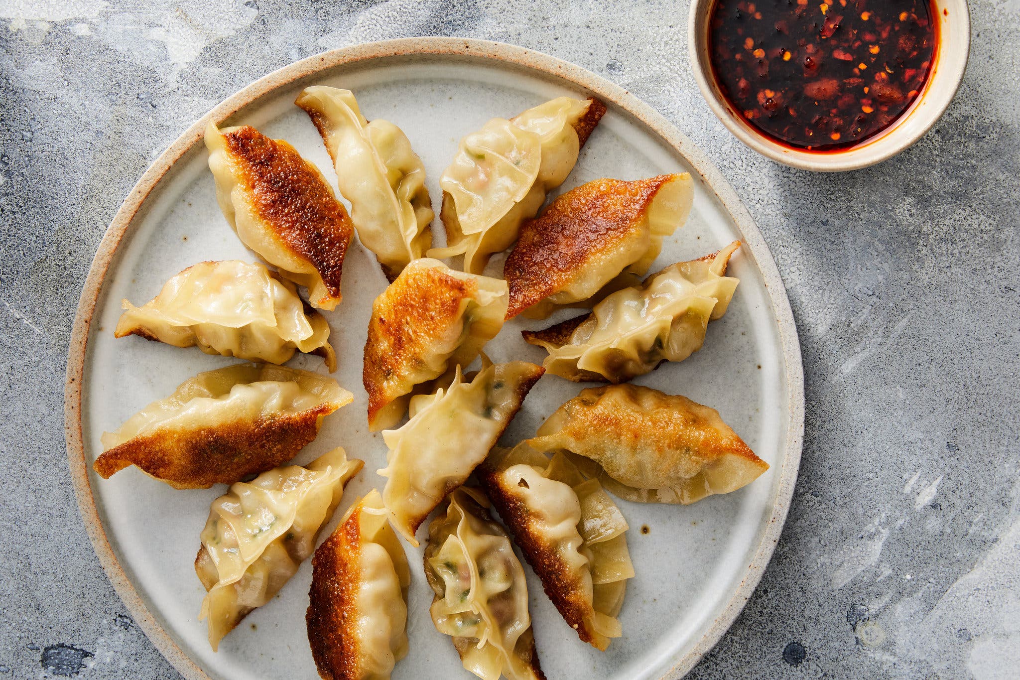 An overhead image of a gray pottery plate topped with dumplings, seared golden brown on some sides. A bowl of dark red sauce specked with seeds in a small white bowl peeks into the frame.