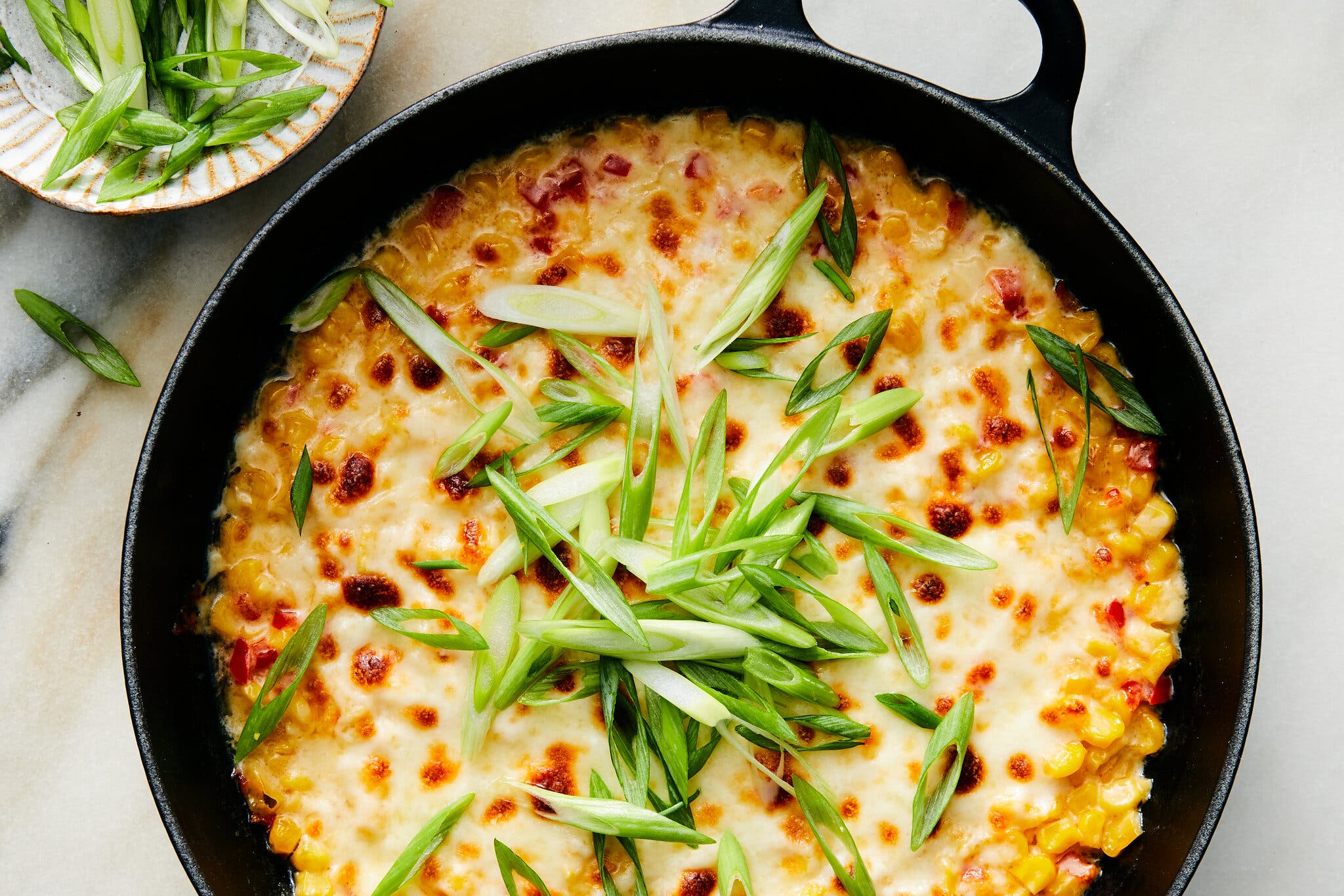 An overhead image of a cast-iron skillet filled with corn kernels topped with melted and browned white cheese and sliced scallion greens. 