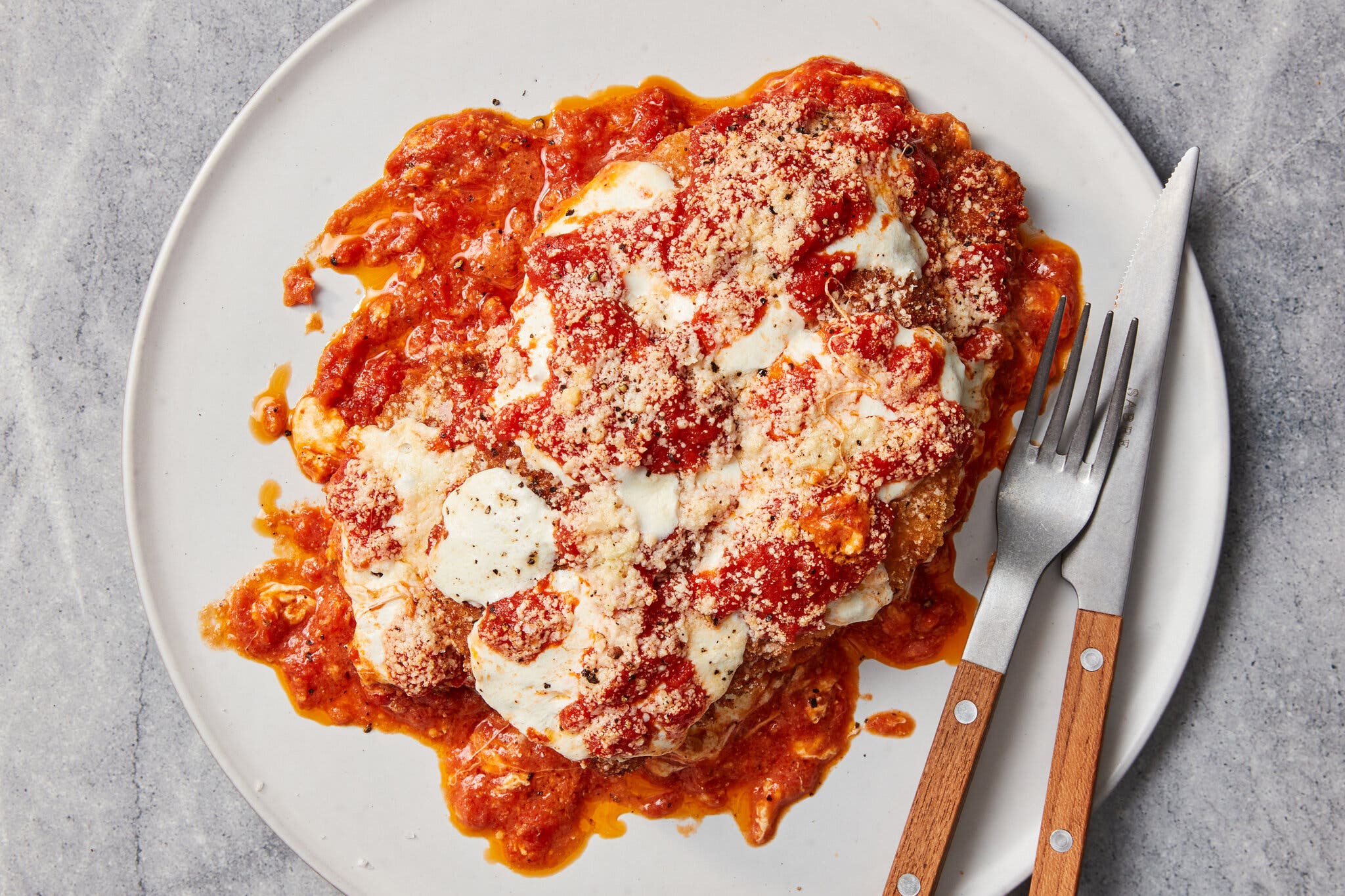 An overhead shot of a white plate with a large boneless chicken breast doused in tomato sauce, melted mozzarella and grated Parmesan. 