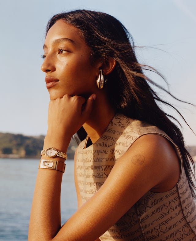 a person rests their chin on their hand wearing a sleeveless outfit with a patterned design and several accessories