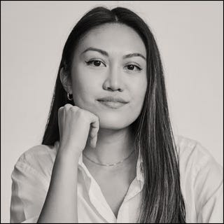 a person with long hair is sitting and resting their chin on their hand wearing a white shirt and a necklace