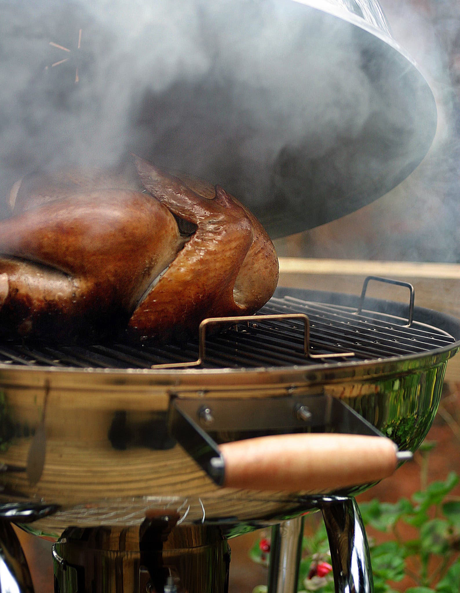 A side image of a golden-brown turkey on a grill. The lid is lifted and exposes a good amount of smoke.