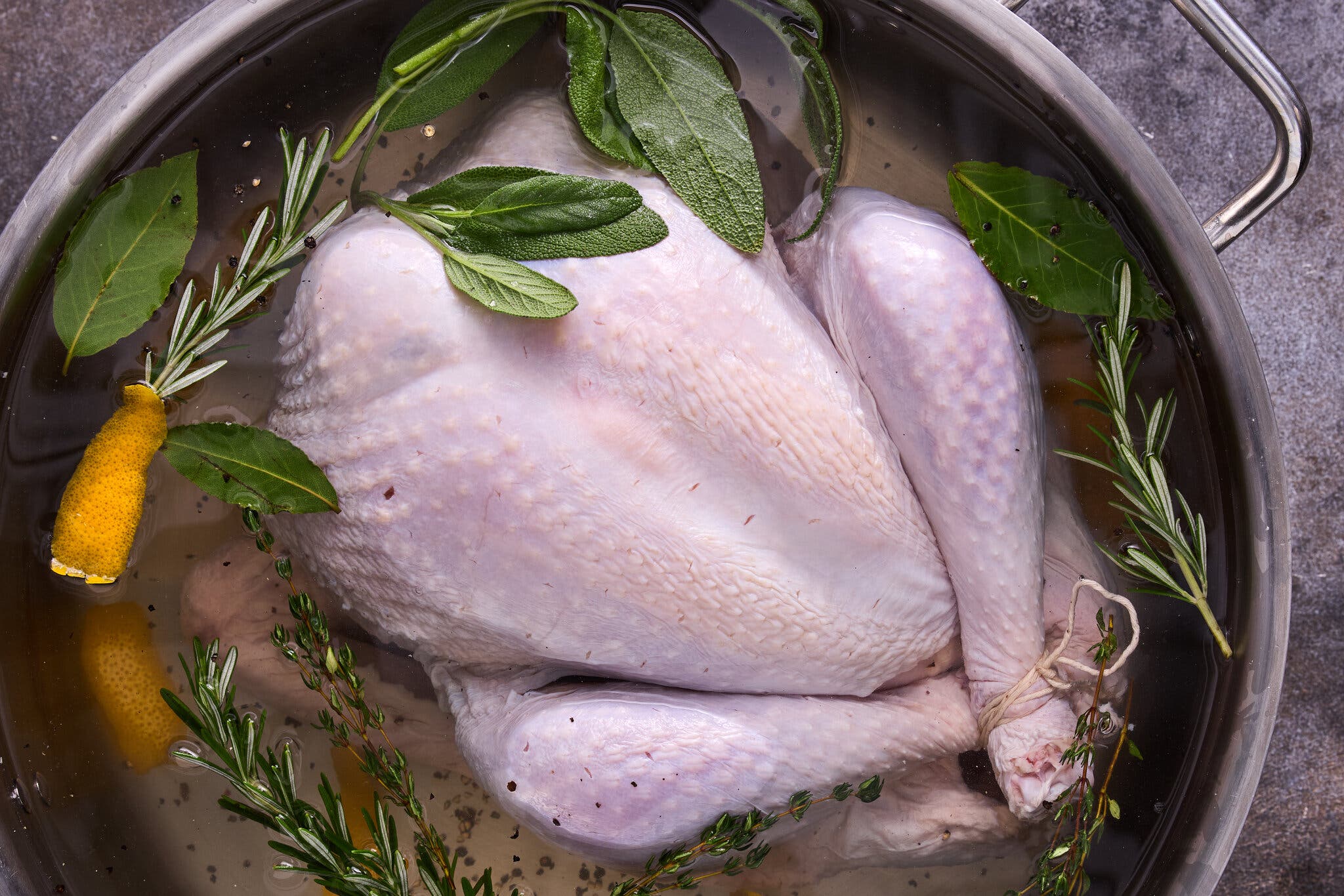 An overhead image of an uncooked turkey in a pot of water surrounded by herbs.