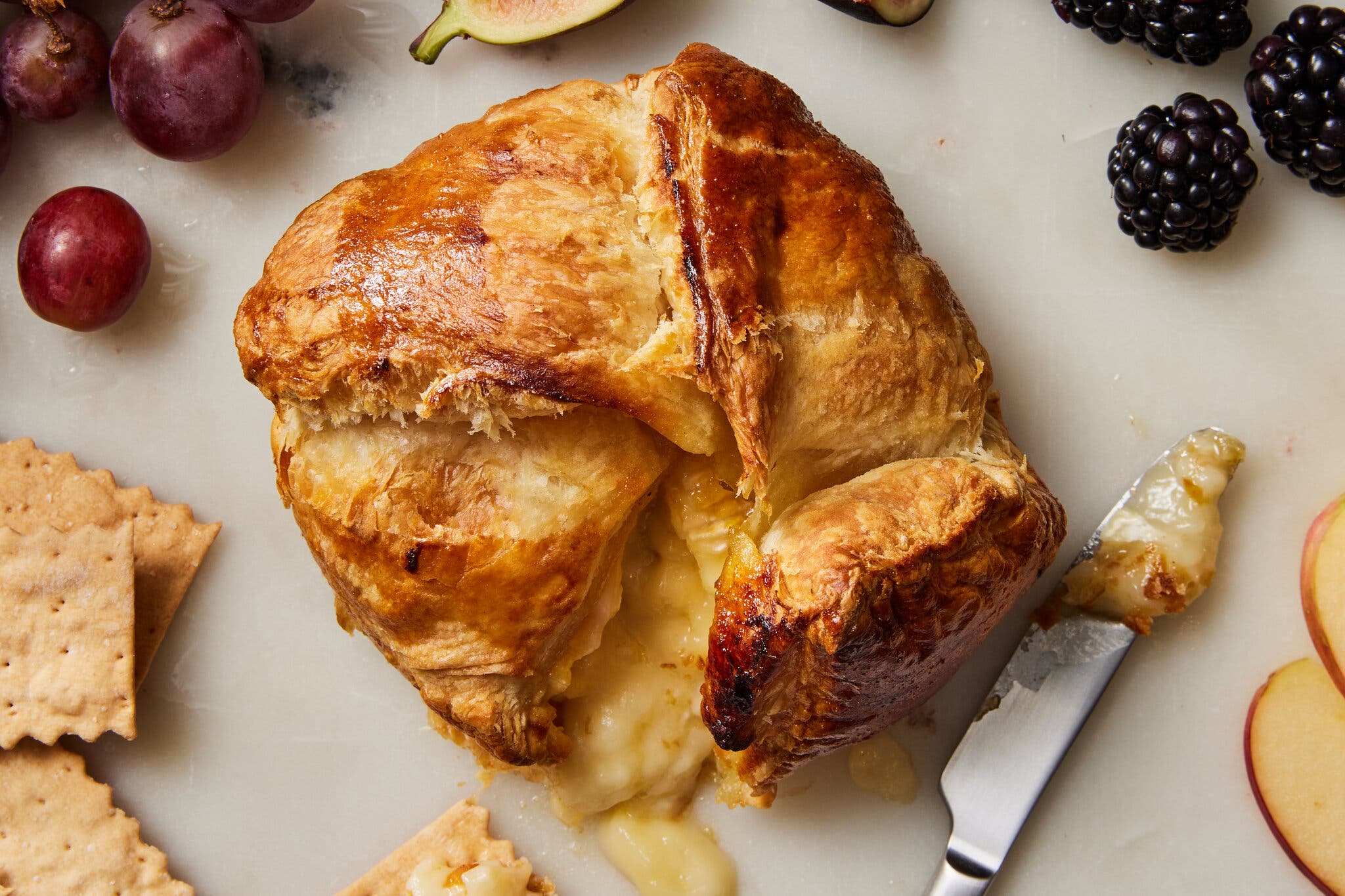 A folded package of baked Brie has been cut open to show the oozing cheese inside. It’s surrounded by crackers, grapes and blackberries.