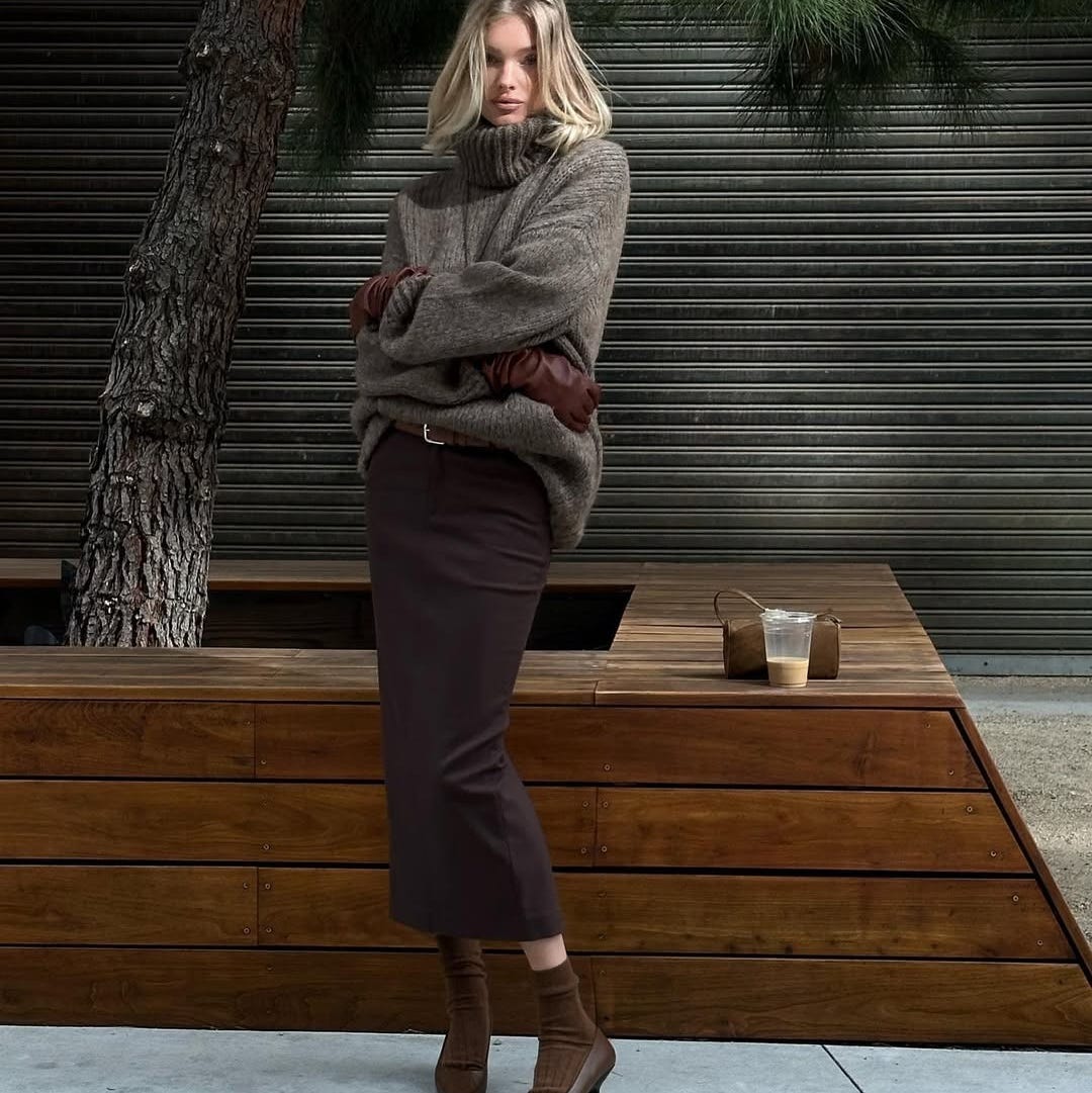 a person stands with arms crossed wearing a chunky sweater a long skirt and heeled boots next to a wooden structure