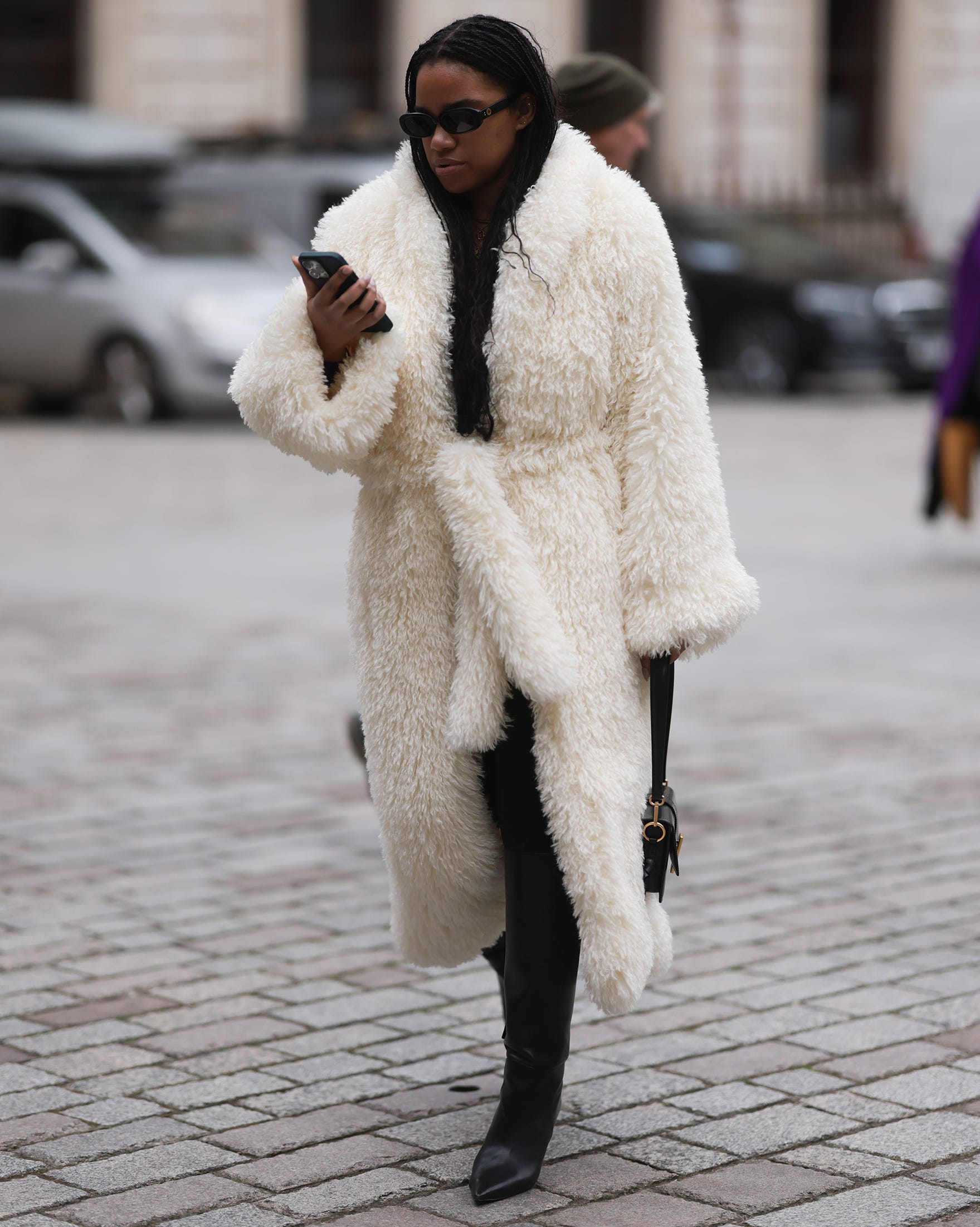 person wearing a long fluffy cream coat and black boots holding a phone while walking on a cobblestone street