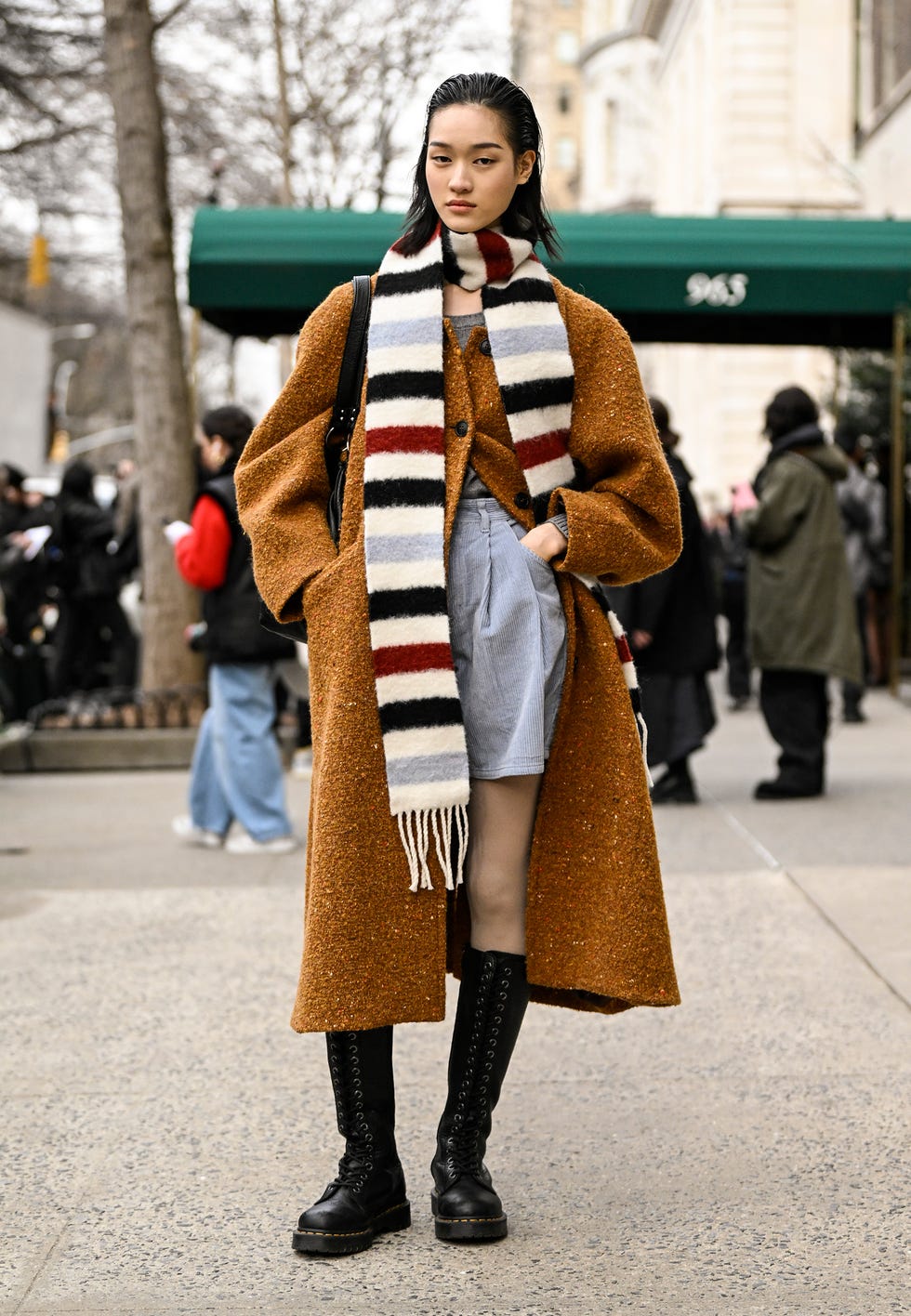 A fashionable individual wearing a brown coat striped scarf and black boots on a city street