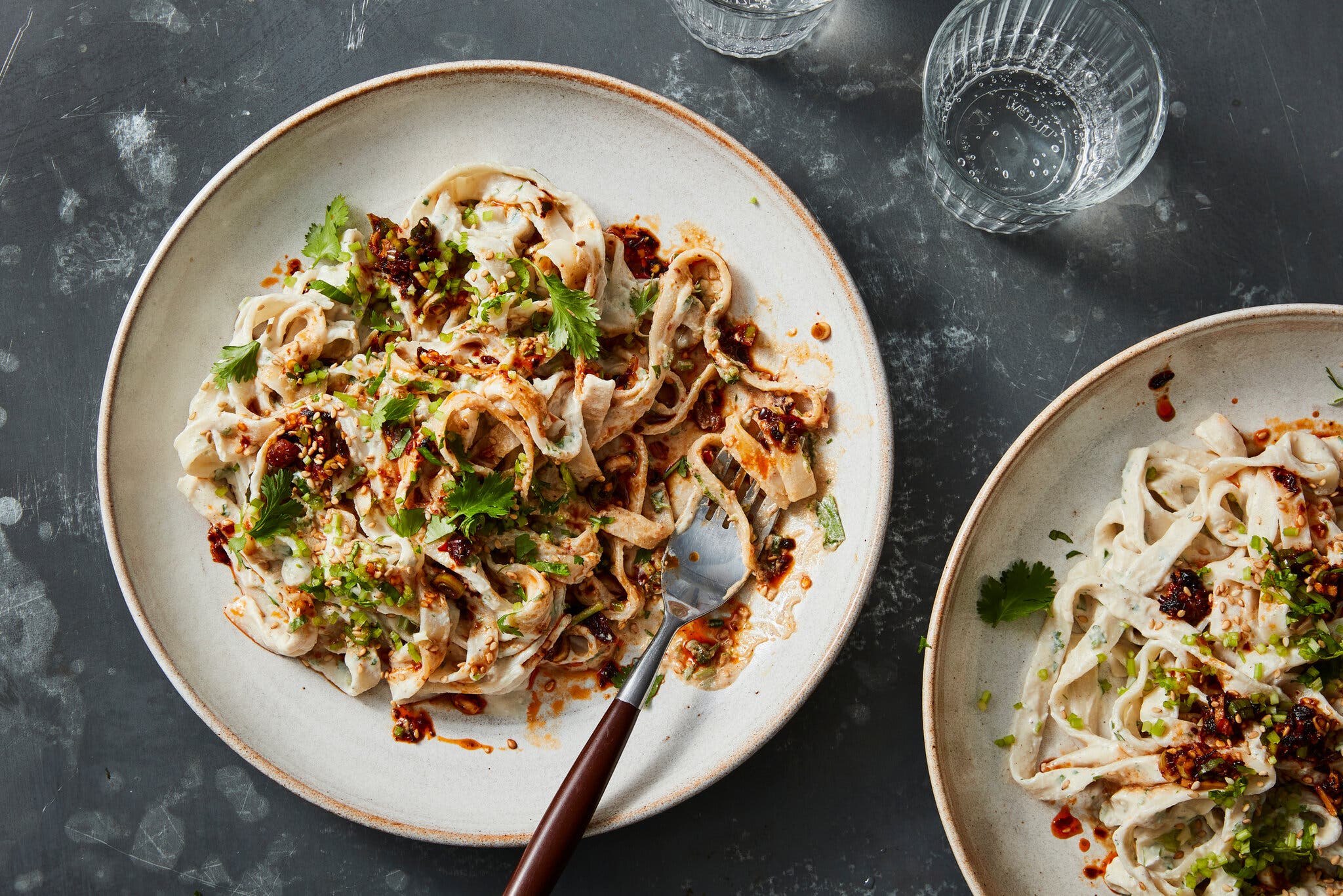 An overhead image of two plates topped with creamy noodles. Chili crisp and herbs are also run throughout.